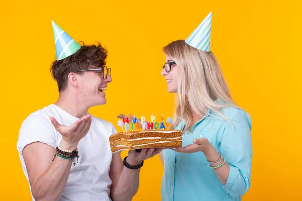 Glad ungt par charmig kille och söt flicka i papper hattar göra dåraktigt ansikte och hålla i sina händer en tårta med inskriptionen födelsedag stående på en gul bakgrund. Koncept hälsningar och — Stockfoto