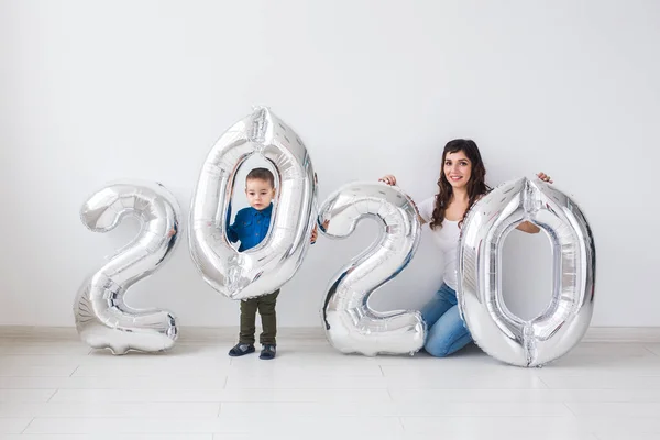 Año nuevo, celebración y concepto de vacaciones - madre e hijo sentados cerca de signo 2020 de globos de plata para el año nuevo en el fondo de la habitación blanca — Foto de Stock