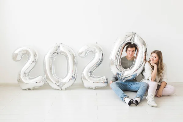 Vacaciones, concepto festivo y de fiesta - Pareja divertida sentada en un piso cerca de globos plateados 2020 sobre fondo blanco. Celebración de Año Nuevo — Foto de Stock