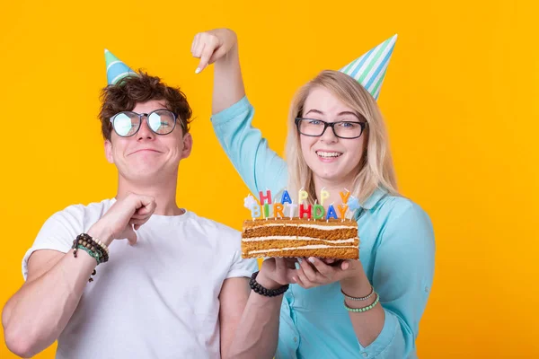 Divertido nerd hombre y mujer están usando vacaciones tapas y gafas celebración de pastel de cumpleaños con velas sobre fondo amarillo — Foto de Stock