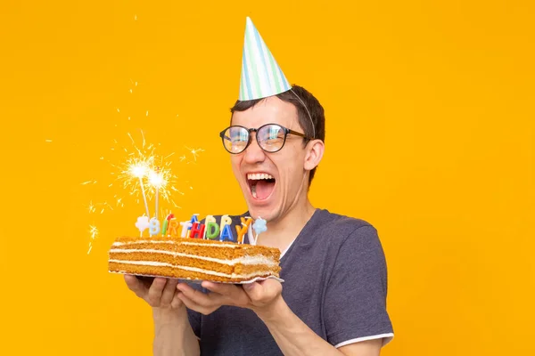 Louco alegre jovem asiático cara com óculos segurando uma vela acesa em suas mãos e um bolo caseiro de parabéns em um fundo amarelo. Conceito de celebração de aniversário e aniversário . — Fotografia de Stock