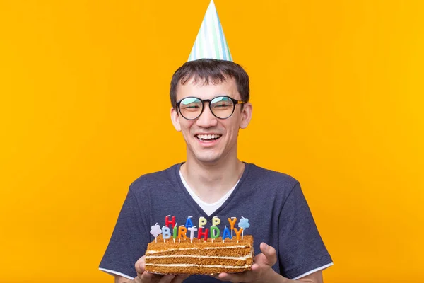 Crazy vidám fiatalember a szemüveg és a papír gratuláló kalap gazdaság sütemények boldog születésnapot állva egy sárga háttér. Jubileumi gratuláció koncepció. — Stock Fotó