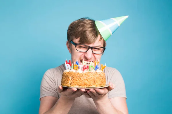 Crazy vicces fiatalember a szemüveg és a papír gratuláló kalap gazdaság sütemények boldog születésnapot állva a kék háttér copyspace — Stock Fotó
