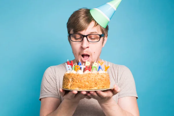 Verrückter lustiger junger Mann mit Brille und Gratulationshüten aus Papier mit Kuchen zum Geburtstag auf blauem Hintergrund stehend. — Stockfoto