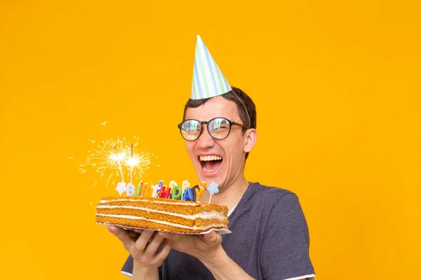 キャップと燃えるキャンドルと黄色の背景にポーズをとる彼の手の中に自家製ケーキを持つ肯定的な面白い若いアジアの男。記念日と誕生日のコンセプト. — ストック写真