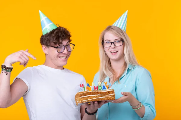 명랑한 젊은 부부 매력적인 남자와 종이 모자에 귀여운 소녀는 어리석은 얼굴을하고 노란색 배경에 서 비문 생일과 함께 자신의 손에 케이크를 개최합니다. 컨셉 인사말 및 — 스톡 사진
