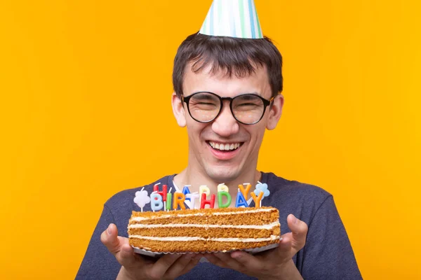 Szalony wesoły młody azjatycki facet z okularami trzyma płonącej świecy w dłoniach i gratulacyjny domowej roboty ciasto na żółtym tle. Koncepcja obchodów urodzin i rocznicy. — Zdjęcie stockowe
