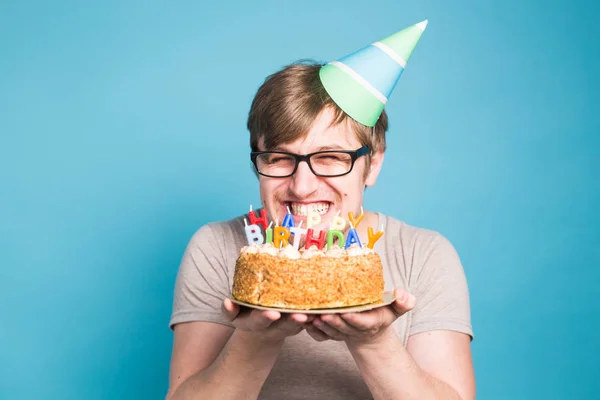 Verrückter lustiger junger Mann mit Brille und Gratulationshüten aus Papier mit Kuchen zum Geburtstag auf blauem Hintergrund mit Kopierraum — Stockfoto