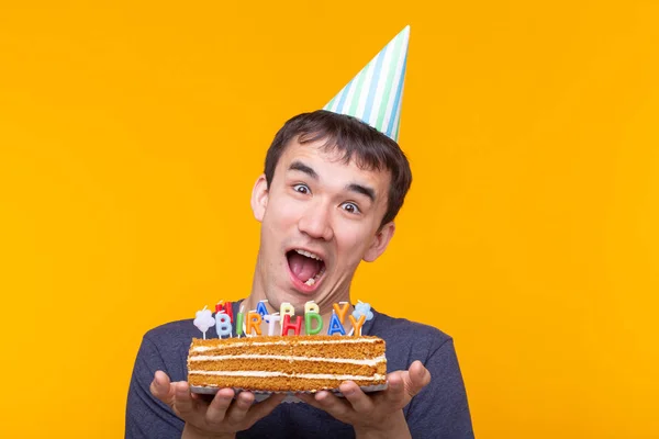 Ein verrückter, fröhlicher junger Mann mit Gläsern und Gratulationsmützen aus Papier, auf gelbem Grund stehend, mit Kuchen zum Geburtstag. Glückwunsch zum Jubiläum. — Stockfoto