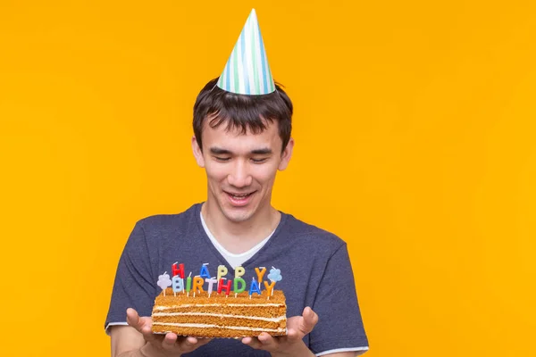 Gözlük ve kağıt tebrik şapkadeli neşeli genç adam kopya alanı ile sarı bir arka plan üzerinde duran pastalar mutlu doğum günü tutan. Jubilee tebrikler kavramı. — Stok fotoğraf