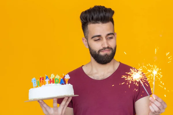 Pozitív fiatal férfi, aki egy boldog születésnapi torta és két égő bengáli lámpák pózol a sárga háttér. — Stock Fotó