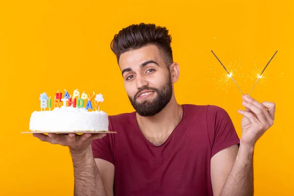 Pozitív vicces fiatal fickó egy sapkát, és egy égő gyertyát, és egy házi készítésű sütemény a kezében pózol egy sárga háttér. Évforduló-és születésnapi koncepció. — Stock Fotó