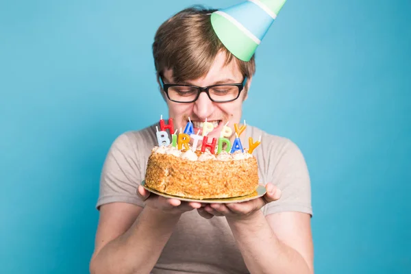Zabawny mężczyzna w papier gratulacyjny kapelusz próbuje ugryźć ciasto z Happy Birthday świece stoją na niebieskim tle. — Zdjęcie stockowe