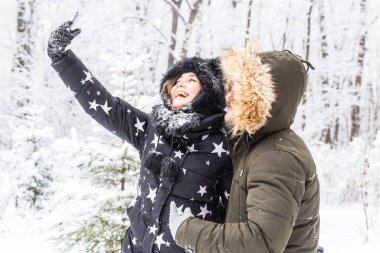 Sezon, aşk, teknoloji ve eğlence konsepti. Mutlu çift kış arkaplanı boyunca akıllı telefonla selfie çekiyor.