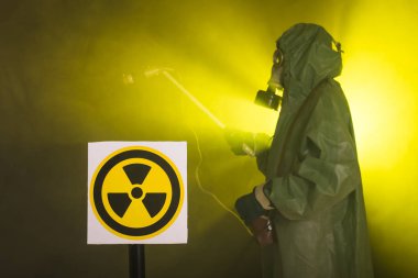 Radyasyon, kirlilik ve tehlike kavramı - koruyucu giysiler içinde bir adam ve karanlık arka planda bir gaz maskesi
