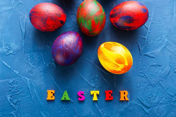 Mnohobarevná dřevěná písmena tvořící slova veselé velikonoce a dekorativní barevná vejce na modrém pozadí. Horní pohled. — Stock fotografie