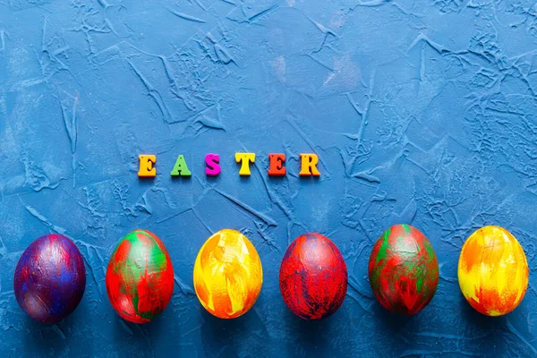 Letras de madera multicolores que componen las palabras feliz Pascua y huevos decorativos de colores sobre un fondo azul. Vista superior . —  Fotos de Stock