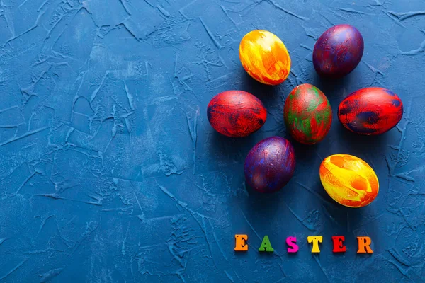 Mnohobarevná dřevěná písmena tvořící slova veselé velikonoce a dekorativní barevná vejce na modrém pozadí s kopírovacím prostorem. Horní pohled. — Stock fotografie