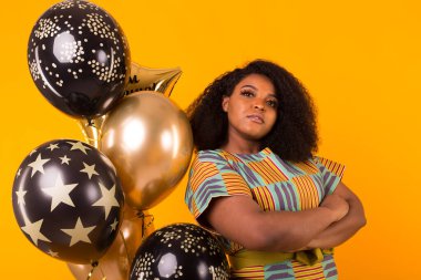 Holidays, party and fun concept - Portrait of smiling young African-American young woman looking sweet on yellow background holding balloons.