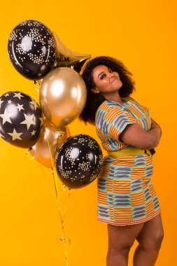 Holidays, party and fun concept - Portrait of smiling young African-American young woman looking sweet on yellow background holding balloons.
