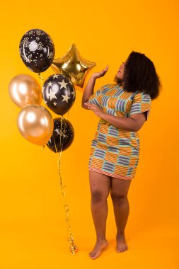 Holidays, party and fun concept - Portrait of smiling young African-American young woman looking sweet on yellow background holding balloons.