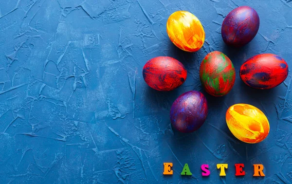 Mnohobarevná dřevěná písmena tvořící slova veselé velikonoce a dekorativní barevná vejce na modrém pozadí s kopírovacím prostorem. — Stock fotografie