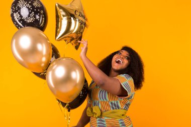 Holidays, party and fun concept - Portrait of smiling young African-American young woman looking sweet on yellow background holding balloons.