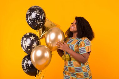Holidays, party and fun concept - Portrait of smiling young African-American young woman looking sweet on yellow background holding balloons.
