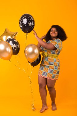 Tatiller, doğum günü partisi ve eğlenceli konsept - gülümseyen Afro-Amerikalı genç bir kadının portresi sarı arka planda balon tutarken tatlı görünüyor.