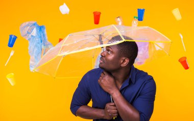 Dünya Çevre Günü, plastik geri dönüşüm sorunu ve çevresel felaket konsepti - sarı arka planda çöplere bakan afro-amerikan erkeği.