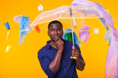 Çöp, plastik geri dönüşüm, kirlilik ve çevre konsepti sorunu - sarı arka planda çöplü dalgın Afrikalı Amerikalı adam.