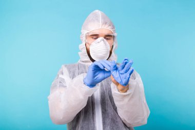 Beyaz dezenfekte kıyafetli bir adam mavi arka planda izole edilmiş tıbbi eldiven giyiyor. Coronavirus, covid-19 ve pandemik konsept.