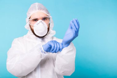 Beyaz dezenfekte kıyafetli bir adam mavi arka planda izole edilmiş tıbbi eldiven giyiyor. Coronavirus, covid-19 ve pandemik konsept.