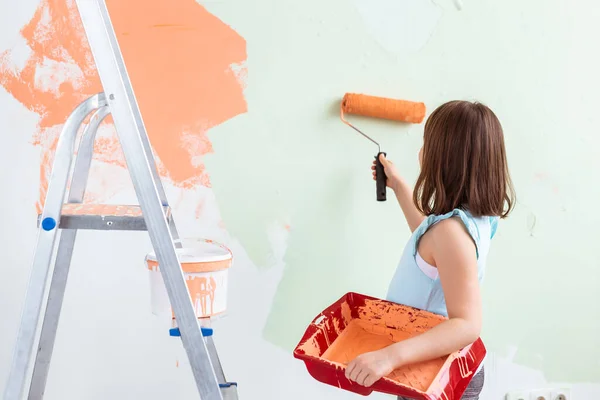 Kid standing with paint roller in hand. She is painting the wall, rear view. Redecoration, repair and repaint concept. — Stock Photo, Image