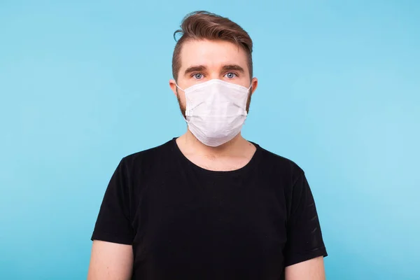 Beau jeune homme en t-shirt noir portant un masque médical sur fond bleu isolé. Covid-19, concept de coronavirus et de virus . — Photo