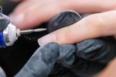 Profesyonel güzellik uzmanlarının elleri müşterinin tırnaklarında elektrikli matkapla çalışıyor. Yapay tırnakların uygulanması prosedürü. Aletli manikürcünün elleri. Eldivenli manikürcü tırnak etlerini çıkarıyor.