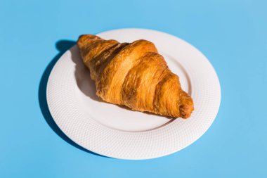 Fırın ürünleri beyaz tabakta pişmiş kruvasan. Mavi arkaplan, en üstteki yakın plan. Pop sanat tarzı. Yaz gölgeleri. Lezzetli ve yemek konsepti.