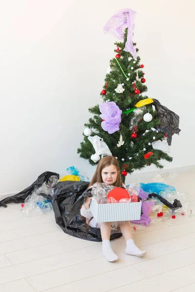 Schockiertes kleines Mädchen blickt mit geöffneten Augen und besorgtem Gesichtsausdruck und hält Schachtel mit verschiedenen Plastikabfällen über dem Christbaumhintergrund. — Stockfoto