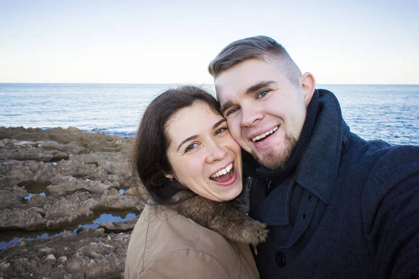 Retrato de jovem casal bonito tirando foto selfie em um smartphone com mar e céu escuro nublado no fundo. Temporada fria e conceito de viagem . — Fotografia de Stock