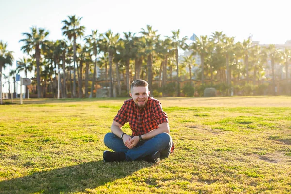 公園で草の上に座っているハンサムな若い笑顔の男。街のコンセプトで夏. — ストック写真