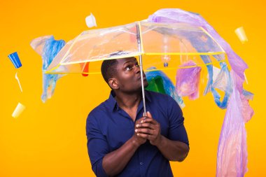 Dünya Çevre Günü, plastik geri dönüşüm sorunu ve çevresel felaket konsepti - sarı arka planda çöplere bakan afro-amerikan erkeği.