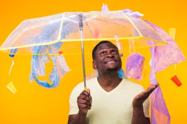 Dünya Çevre Günü, plastik geri dönüşüm sorunu ve çevresel felaket konsepti - sarı arka planda çöplere bakan afro-amerikan erkeği.