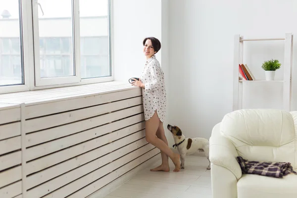 Nyugodt és barátságos. Gyönyörű fiatal nő csésze tea és kutya áll az ablak mellett otthon — Stock Fotó