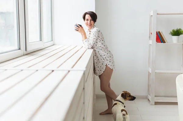 落ち着いて居心地の良い。家の窓の近くに立っているお茶と犬のカップを持つ美しい若い女性 — ストック写真