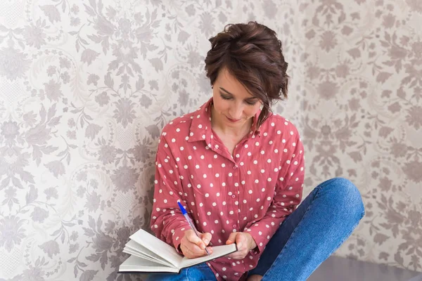 Mladá brunetka odpočívající doma píše zápisník. Karanténa, izolace, pandemický svět koronaviru. Zůstaň doma.. — Stock fotografie