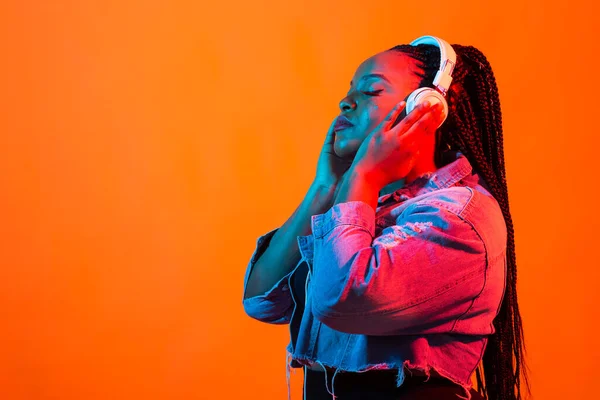 African american young woman listening to music online dancing and singing with headphones, neon light and copy space. Music and technology concept.
