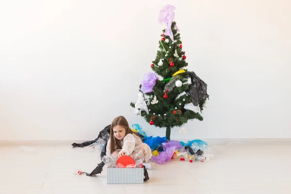 Niña impactada mira con los ojos abiertos y expresión preocupada, caja de espera con varios residuos de plástico sobre el fondo del árbol de Navidad . — Foto de Stock