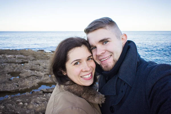 Retrato de jovem casal bonito tirando foto selfie em um smartphone com mar e céu escuro nublado no fundo. Temporada fria e conceito de viagem . — Fotografia de Stock