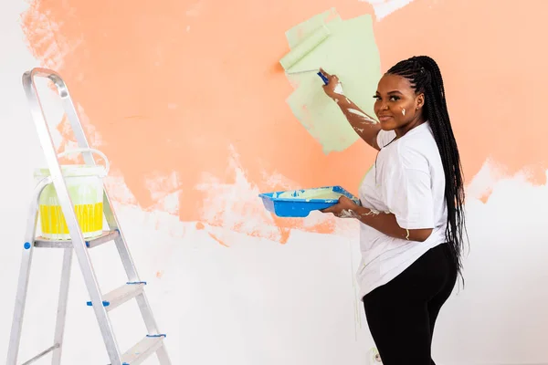 Beautiful african american girl painting the wall with paint roller. Portrait of a young beautiful woman painting wall in her new apartment. Redecoration and renovation concept. — Stock Photo, Image