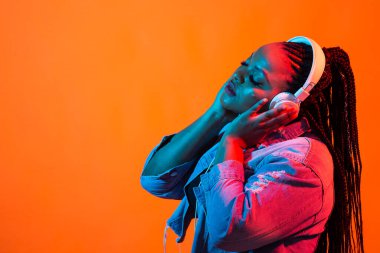 Kulaklıkla müzik dinleyen Afrikalı genç bir kadının Neon portresi. Kopyalama alanı olan turuncu arkaplan.